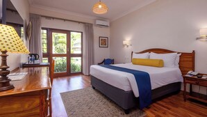 A bedroom with a large bed, a wooden desk, a lamp, a nightstand, and a view of greenery outside.
