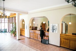 Una recepción con mostrador de madera, una recepcionista y un huésped con maleta.