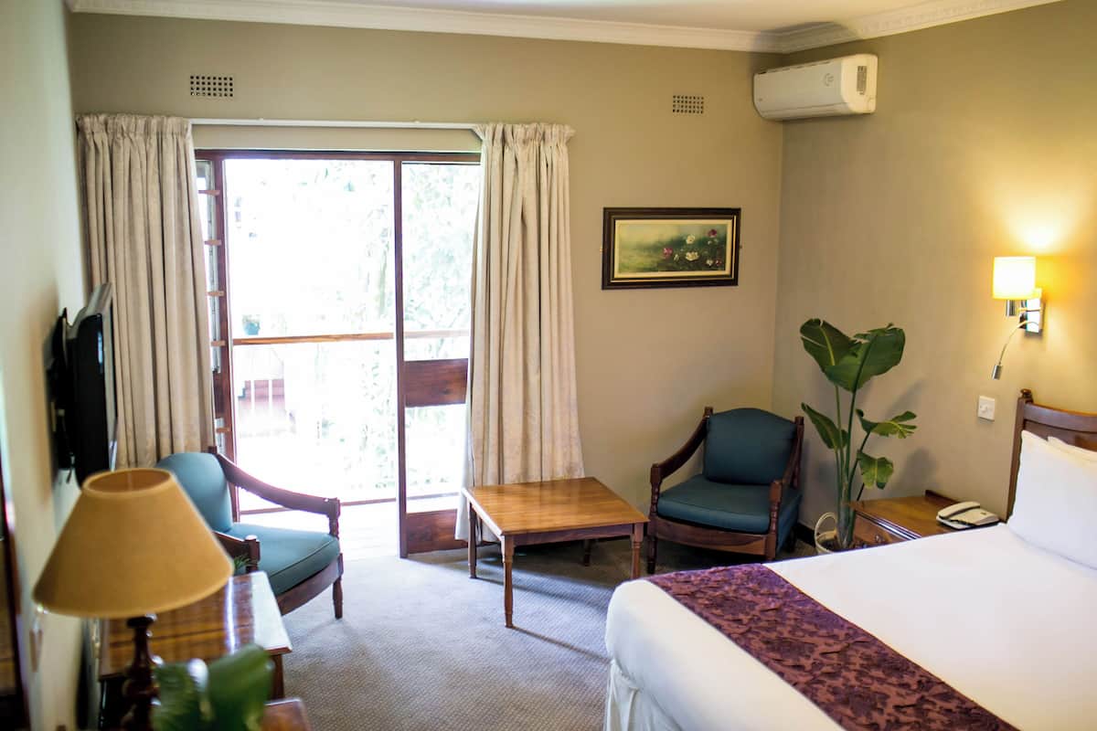 Habitación de hotel con cama, escritorio, dos sillas, una mesa, lámpara, planta, un cuadro y puerta corrediza de vidrio.