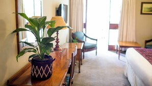 A hotel room with a wooden desk, a potted plant, a chair, a bed, a television, and a window with curtains. - The Bronte Garden Hotel (Harare)