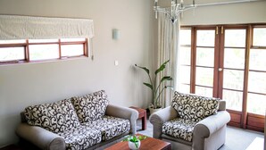 A living room with two patterned sofas, a wooden coffee table, a potted plant, and a large window with curtains. - The Bronte Garden Hotel (Harare)
