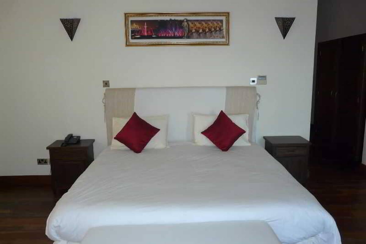 A hotel room with a large bed, two red pillows, a nightstand with a phone, and a painting on the wall.
