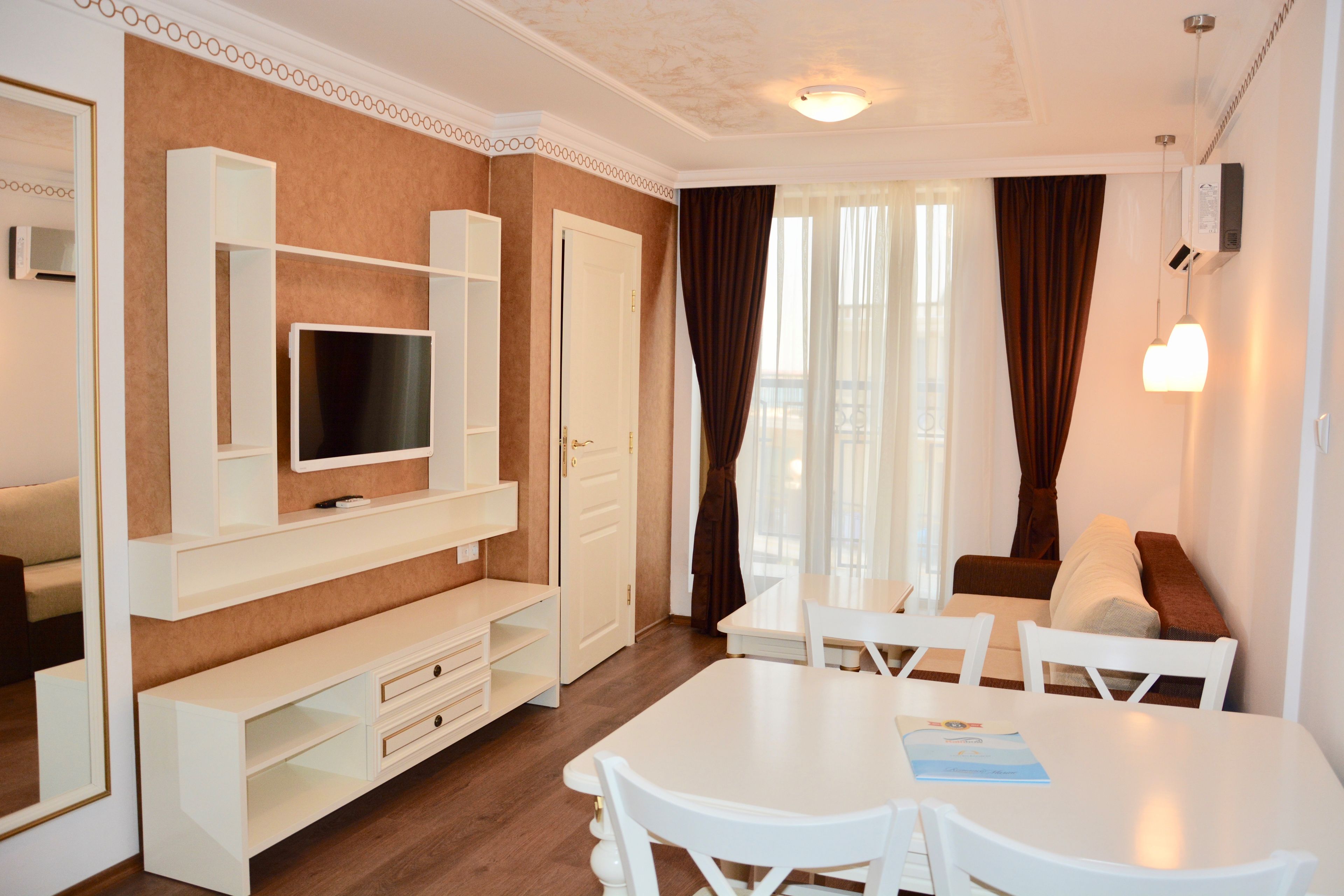 Una sala moderna con televisor de pantalla plana, mobiliario blanco, una mesa de comedor y un ventanal con cortinas.