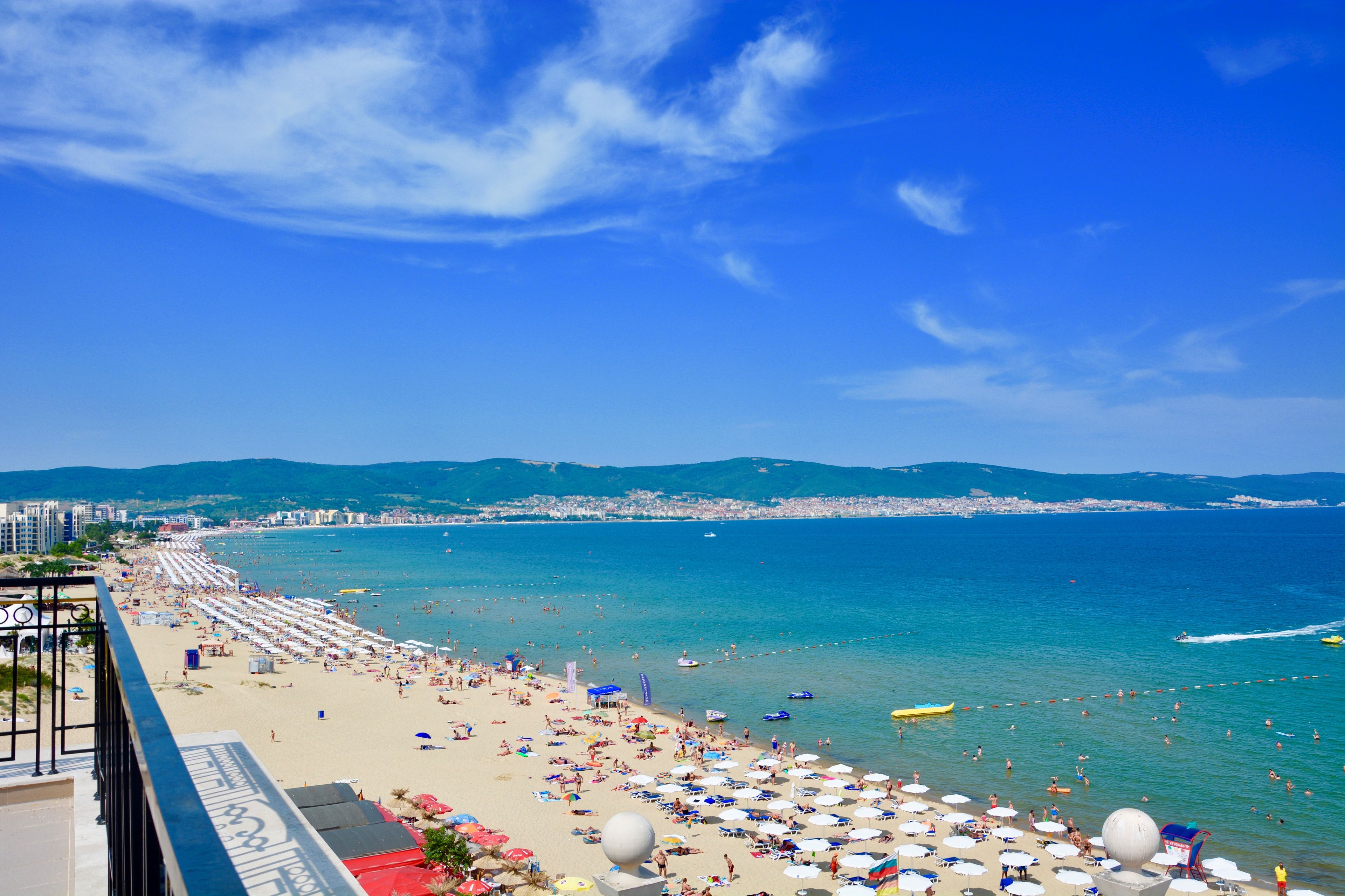 A beach with umbrellas and people, a clear sky, and a view of the sea and distant buildings.