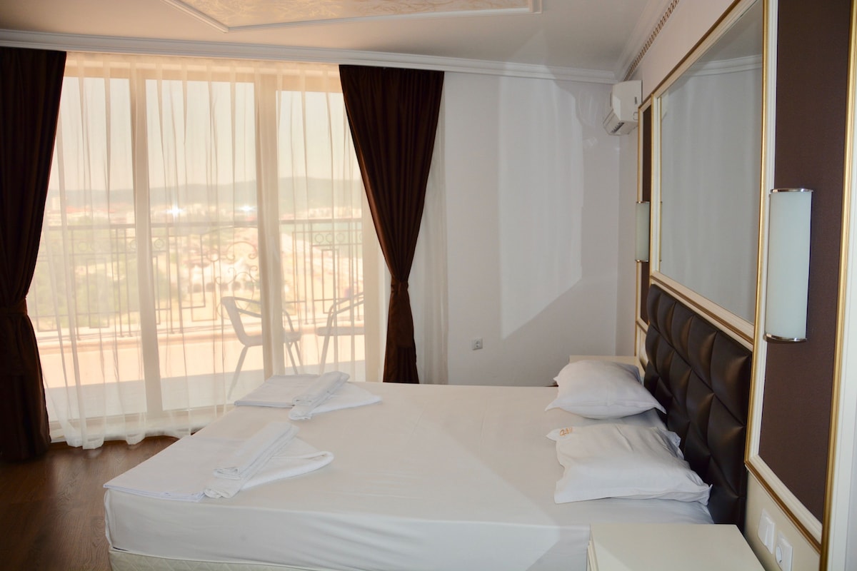 A hotel room with a bed, two pillows, a bedside table, and a window with curtains.