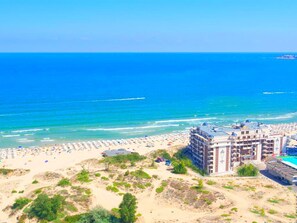 A coastal hotel with multiple floors, a beach area with umbrellas, and a view of the ocean. - Golden Rainbow (Nessebar)