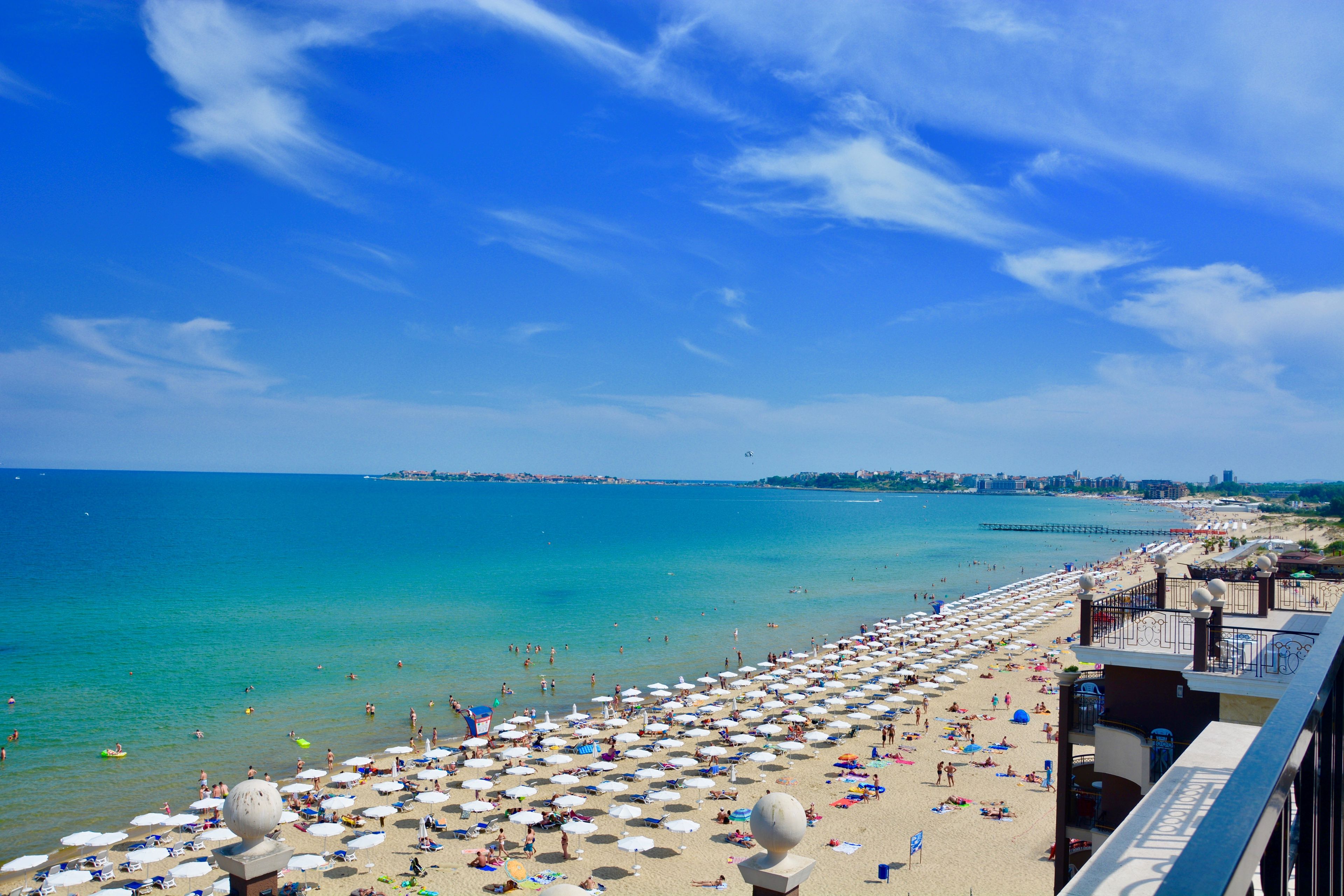 A beach with numerous umbrellas and sun loungers, a clear blue sky, and a view of the sea.