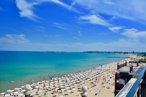 A beach with numerous umbrellas and sun loungers, a clear blue sky, and a view of the sea.