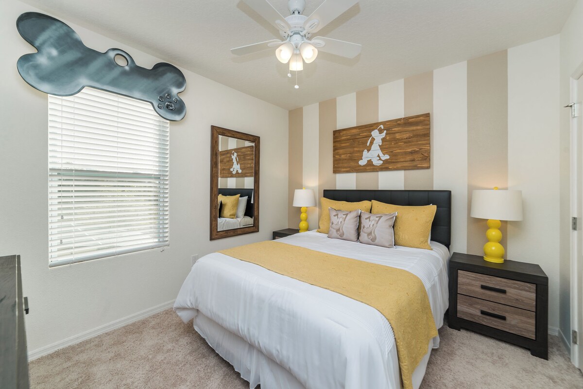 A bedroom with a bed, bedside tables, a ceiling fan, a window with blinds, a mirror, and a wall with a striped pattern.