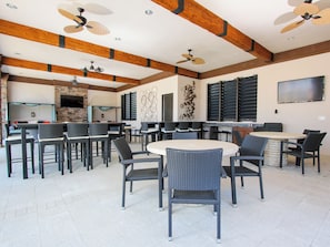 A spacious outdoor dining area with multiple tables and chairs, ceiling fans, and a stone fireplace. - Solterra Resort Homes (Davenport)