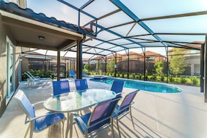 Área de piscina ao ar livre com espaço de estar coberto, mesa e cadeiras.