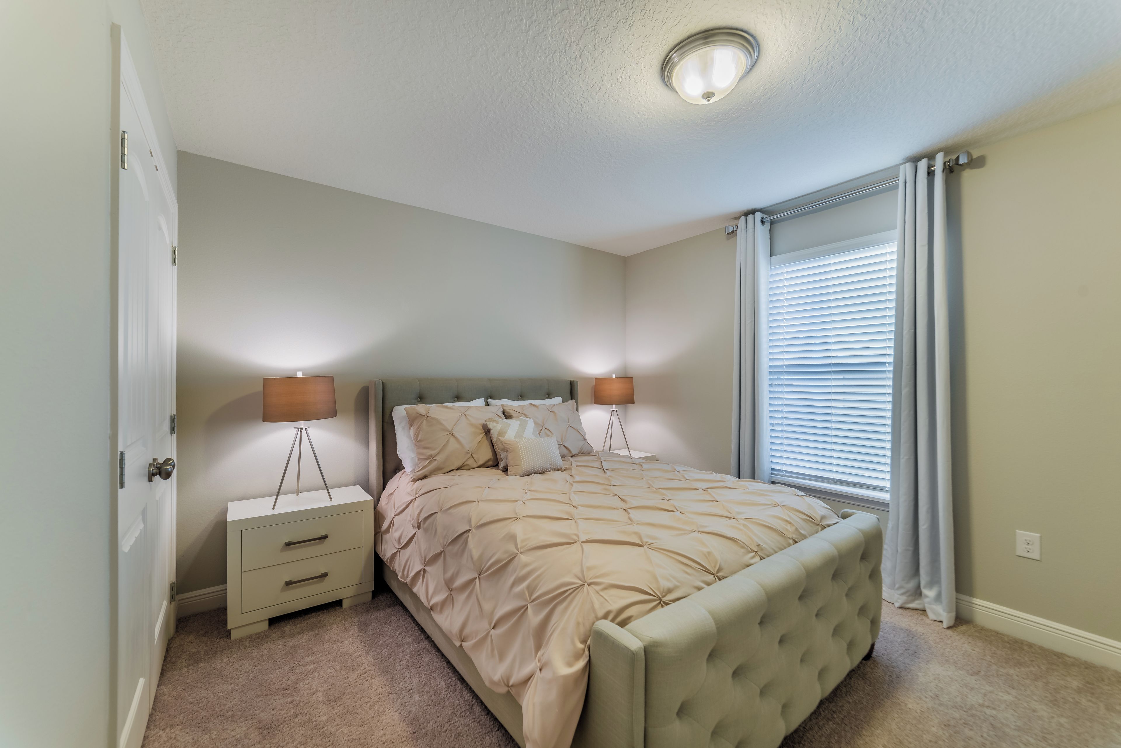 A bedroom with a bed, bedside tables, a lamp, a window with blinds, and a door.