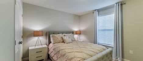 Une chambre à coucher avec un lit, des tables de chevet, une lampe, une fenêtre avec des stores et une porte.