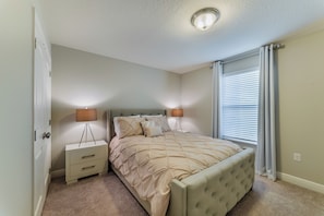 A bedroom with a bed, bedside tables, a lamp, a window with blinds, and a door. - Solterra Resort Homes (Davenport)