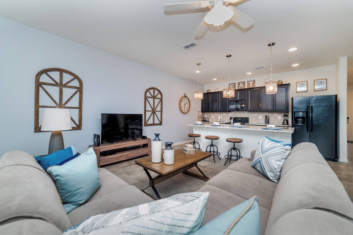 A modern living room with a sofa, coffee table, and TV. A kitchen with bar stools and a refrigerator is visible.