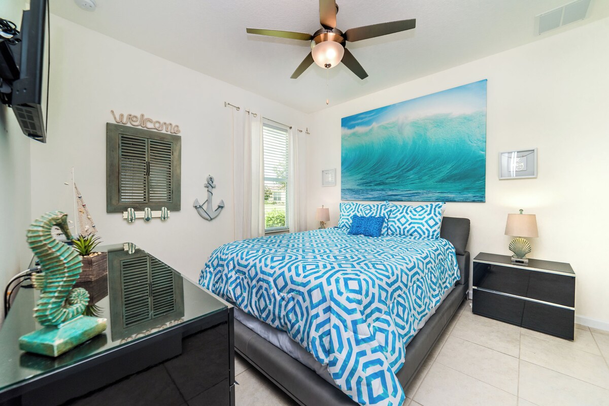 A bedroom with a bed, a ceiling fan, a nightstand, a lamp, a wall-mounted TV, and a window with curtains.