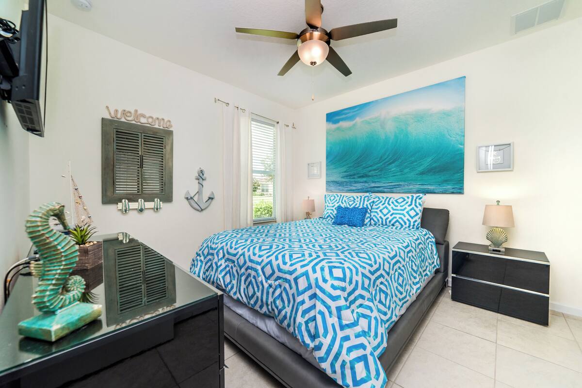 A bedroom with a bed, a ceiling fan, a nightstand, a lamp, a wall-mounted TV, and a window with curtains.
