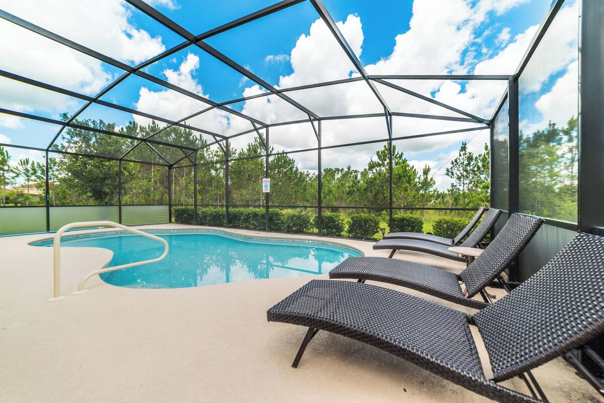 An indoor pool with clear glass walls, lounge chairs, and a view of trees and sky.