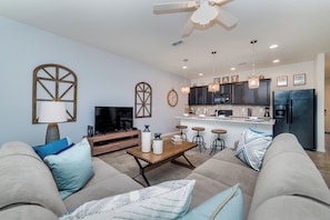 A modern living room with a sofa, coffee table, and TV. A kitchen with bar stools and a refrigerator is visible. - Solterra Resort Homes (Davenport)