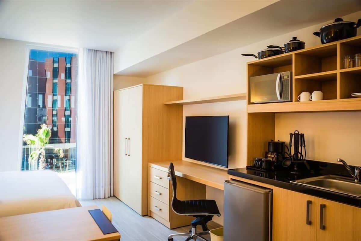 A modern hotel room with a wooden desk, a flat-screen TV, a kitchenette, and a large window with curtains.