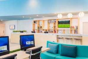 Een moderne hotelentree met een receptiebalie, computers, een zithoek met een blauwgroene bank en een televisie waarop een sportevenement te zien is.