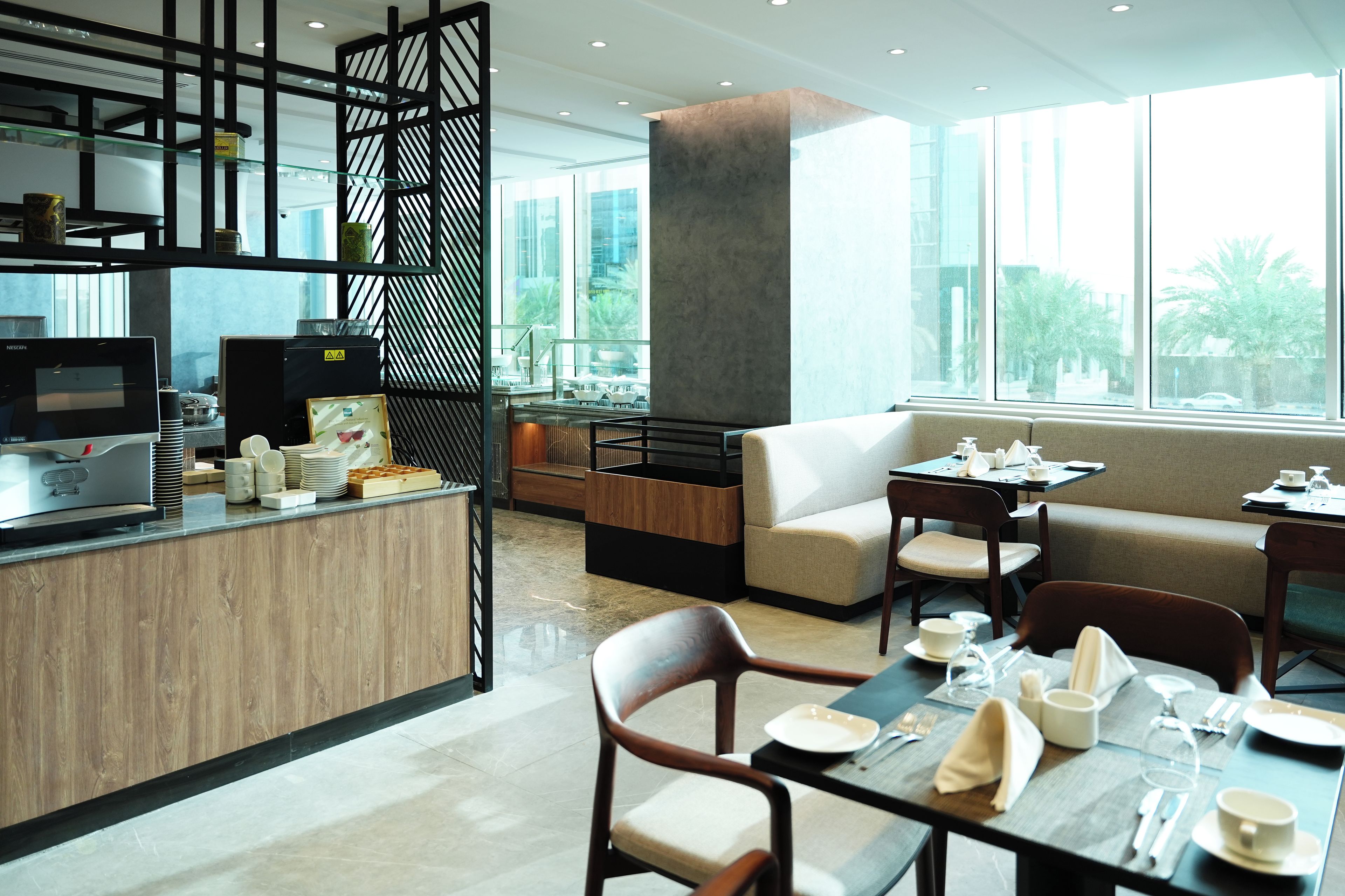 A modern hotel dining area with a wooden counter, seating arrangements, and large windows.