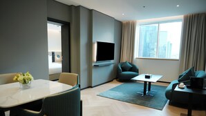 A modern hotel room with a flat-screen TV, a dining table with chairs, a sofa, and a large window with curtains.