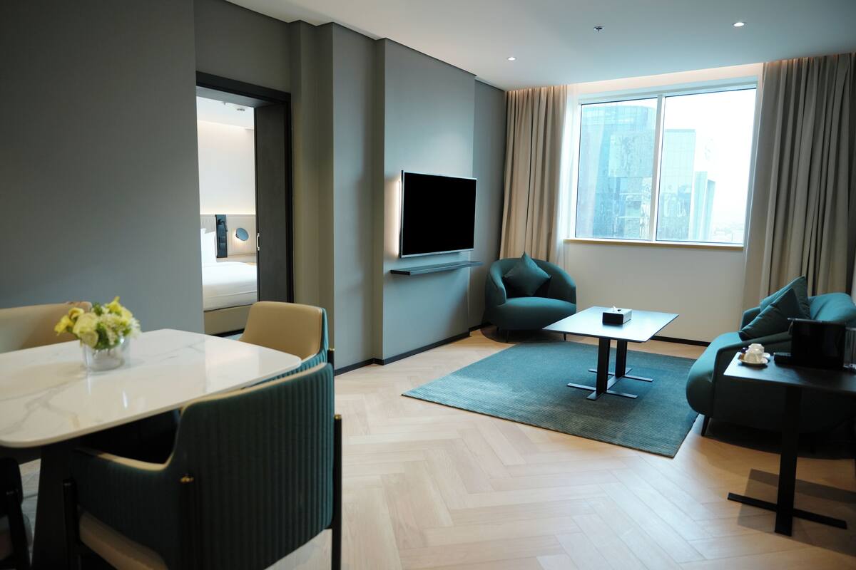 Une chambre d’hôtel moderne dotée d’une télévision à écran plat, d’une table à manger avec des chaises, d’un canapé et d’une grande fenêtre avec des rideaux.