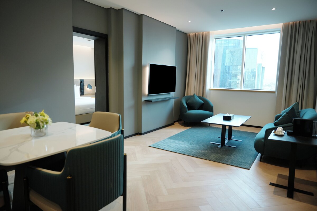 Una habitación de hotel moderna con televisión de pantalla plana, una mesa de comedor con sillas, un sofá y un amplio ventanal con cortinas.
