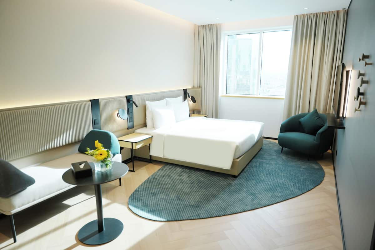 A hotel room with a bed, bedside table, lamp, chair, and a view of the city through the window.