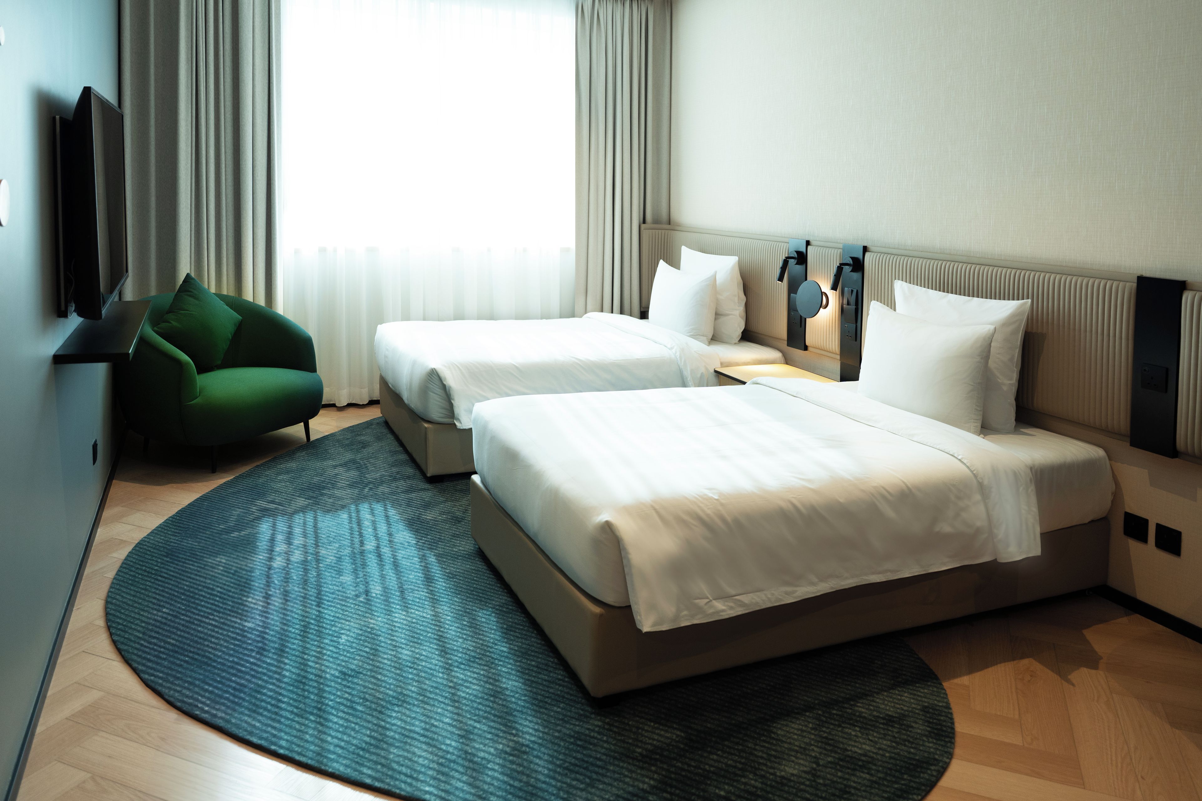 Habitación de hotel con dos camas, un televisor de pantalla plana, un sillón verde y un ventanal con cortinas.
