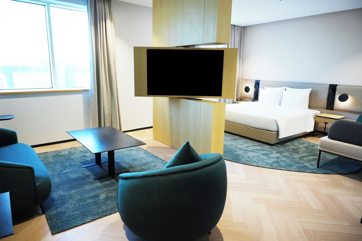 Una habitación de hotel moderna con una cama, televisión montada en la pared, un escritorio y una zona de estar con una silla.