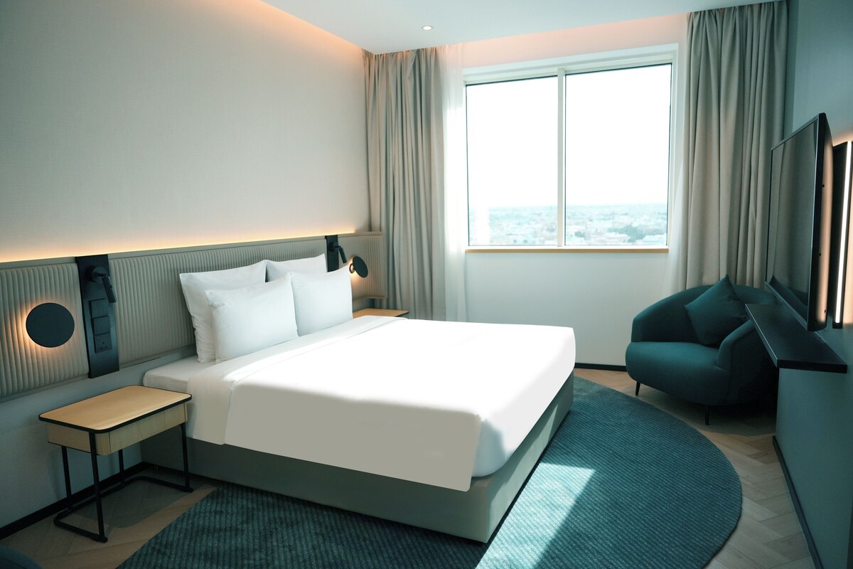 Una habitación de hotel moderna con una cama grande, mesitas de noche, televisión y vistas a la ciudad.