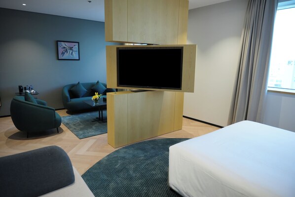 A hotel room with a bed, a sofa, a TV mounted on a wooden cabinet, a small table with a vase, and a window with curtains. - Novotel Riyadh Al Anoud (Al Muzahimiyah)