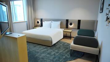 Quarto de hotel com cama, mesas de cabeceira, uma cadeira, televisão e janela com cortinas.