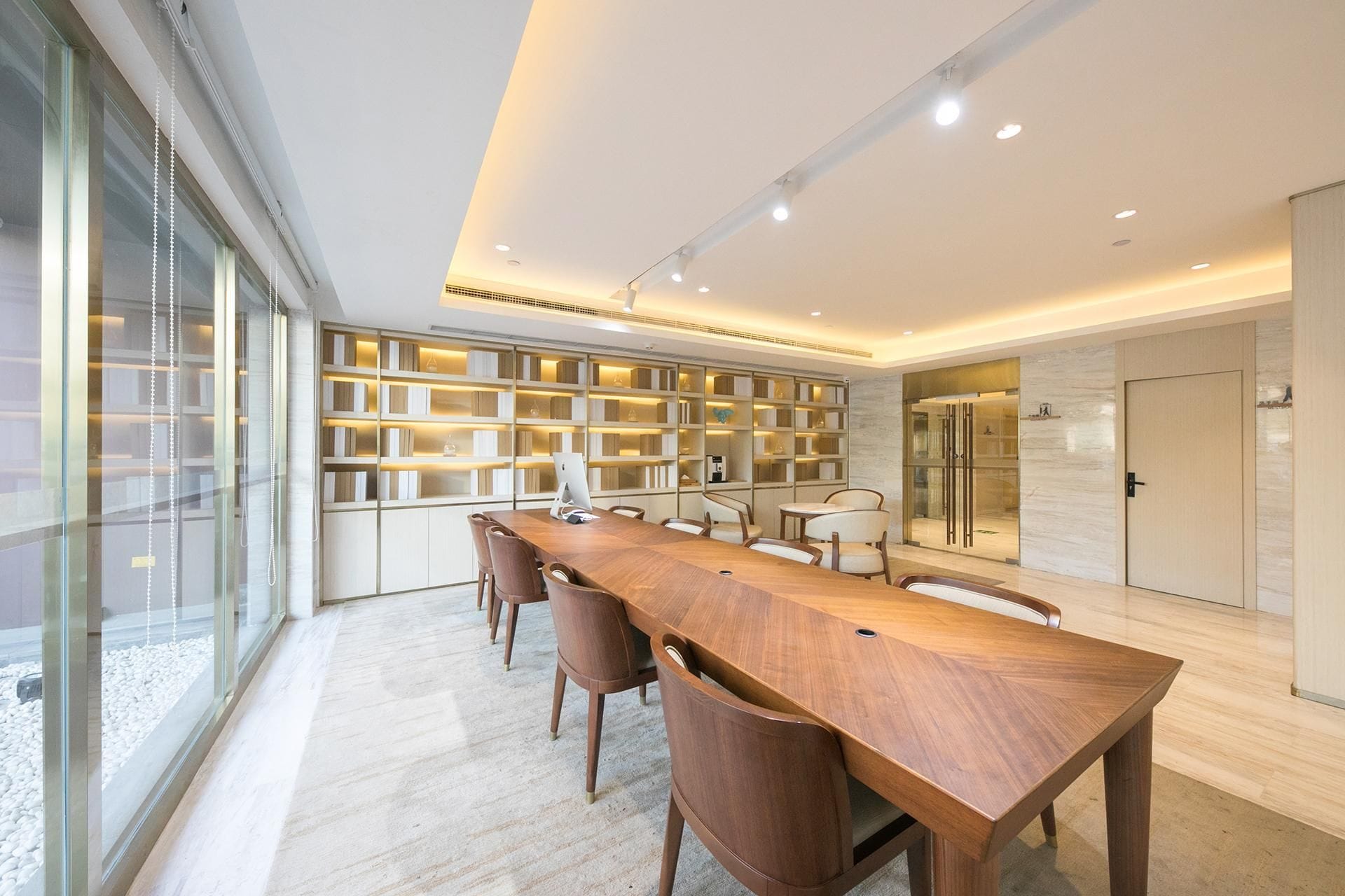 A modern dining area with a long wooden table, several chairs, and a bookshelf filled with books.