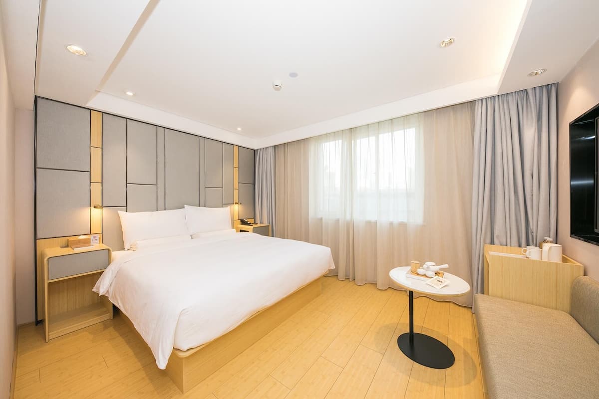 Une chambre d’hôtel moderne avec un grand lit, des tables de chevet, une petite table ronde, un canapé et une grande fenêtre avec des rideaux.