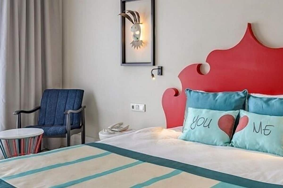 Ein Hotelzimmer mit rotem Kopfteil, einem Bett mit türkis-weißer Bettwäsche, einem blauen Sessel und einem kleinen runden Tisch.