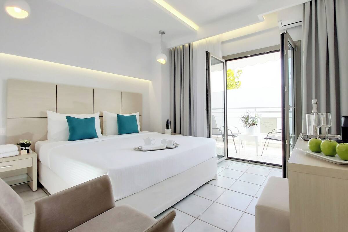 A modern hotel room with a large bed, a desk with a tray of apples, a glass of water, and a bottle.