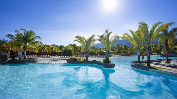 A clear blue pool with palm trees, lounge chairs, and a mountainous backdrop.