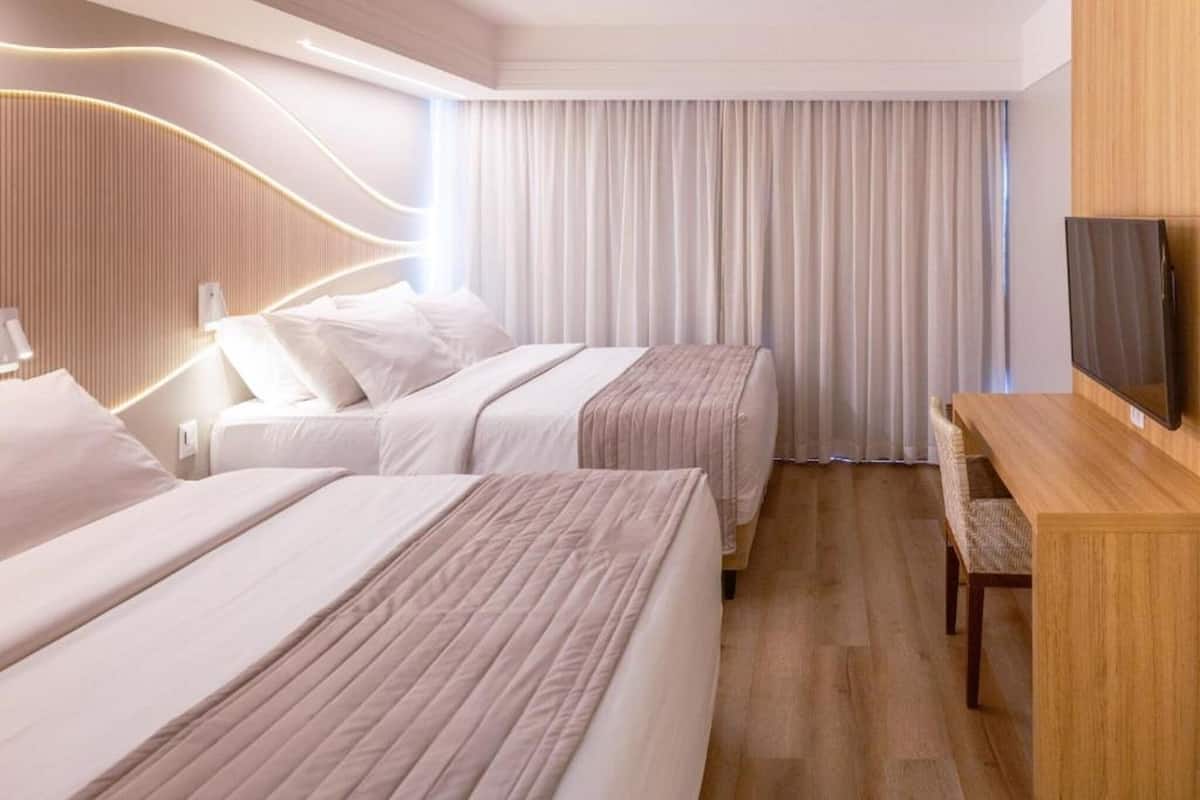 Una habitación de hotel con dos camas, un escritorio de madera, un televisor y un ventanal con cortinas.