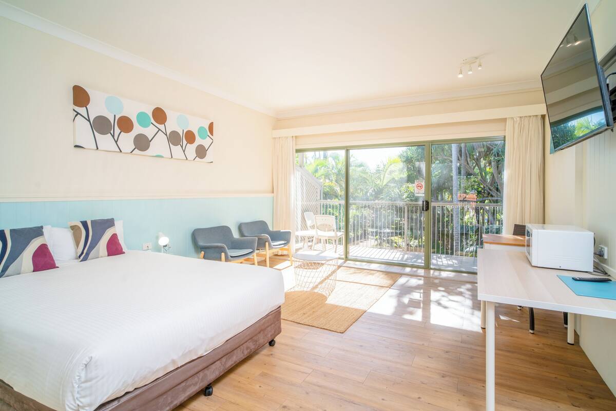 A hotel room with a bed, a desk, a TV, and a balcony with chairs.
