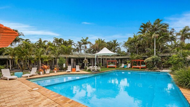 A swimming pool with lounge chairs and a covered seating area, surrounded by palm trees and a building with a tiled roof. - Quality Hotel Ballina (Ballina)