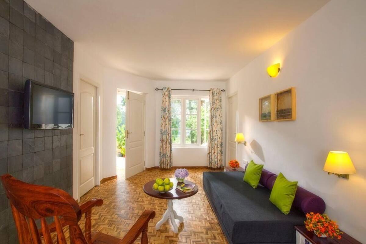 A living room with a sofa, a wooden chair, a small table with fruit, a TV, and a window with curtains.