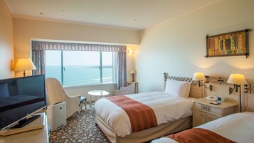 Habitación de hotel con cama, dos mesitas de noche, televisor, sillón, una pequeña mesa y ventana con vista al mar.