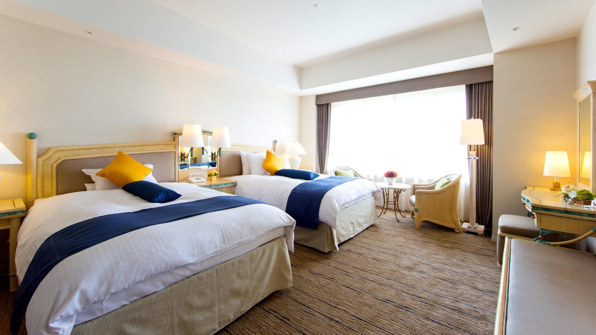 Une chambre d’hôtel avec deux lits, un coin salon comprenant un fauteuil, une petite table et une grande fenêtre.
