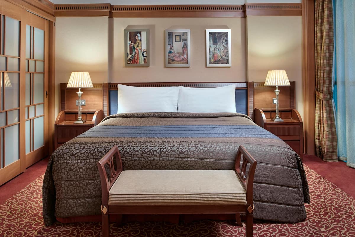 A hotel room with a large bed, two bedside tables with lamps, a chair, and three framed artworks on the wall.