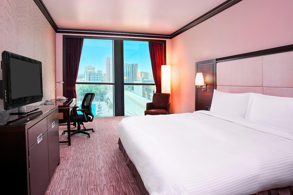A hotel room with a bed, desk, chair, television, and a view of the cityscape.