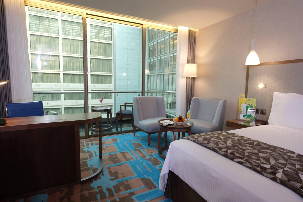 A hotel room with a bed, two armchairs, a desk, a small table with a bowl of fruit, and a window with curtains.