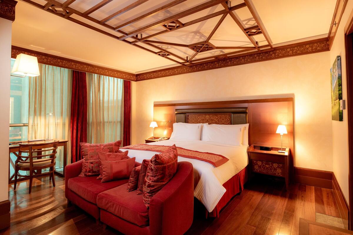 A hotel room with a bed, a red sofa, a wooden desk, and a nightstand with lamps.